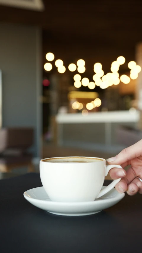 a person holding a cup of coffee on a table, by Arabella Rankin, trending on unsplash, lights on, square, cafe interior, thumbnail