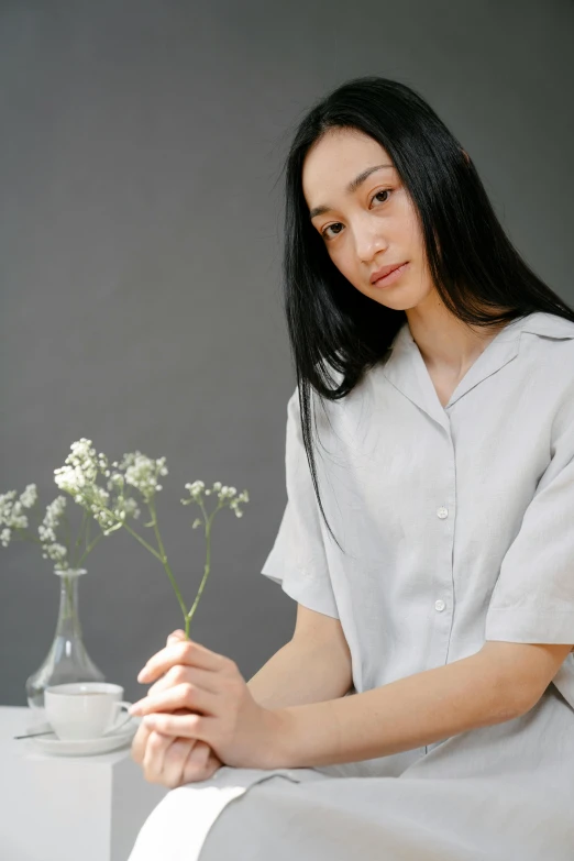 a woman sitting next to a vase of flowers, a portrait, inspired by Ruth Jên, trending on unsplash, minimalism, wearing a linen shirt, light grey, center focus on table, indonesia