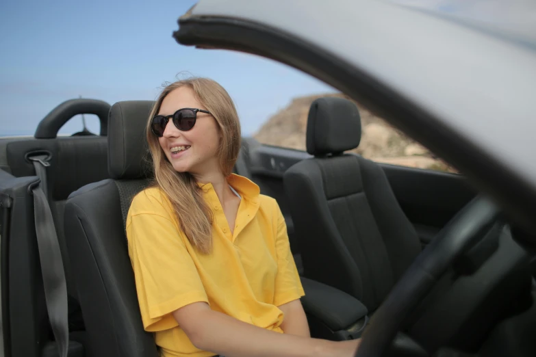 a woman sitting in the driver's seat of a car, inspired by Sydney Carline, wearing polo shirt, yellow sunshine, topdown, wearing shades