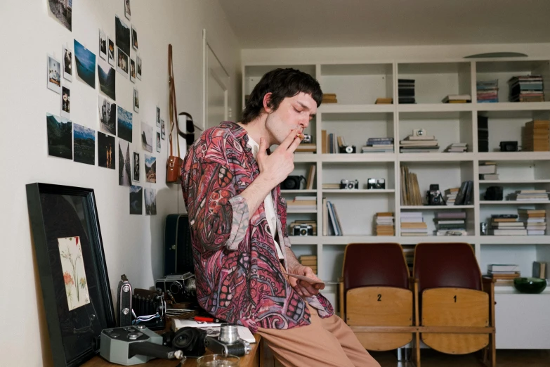 a man sitting on a desk smoking a cigarette, an album cover, by Nina Hamnett, unsplash, in the office, sideburns, paisley, at home