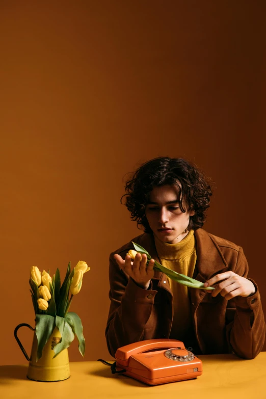 a man sitting at a table with a phone in front of him, an album cover, inspired by Luca Zontini, trending on pexels, carrying flowers, brown curly hair, yellow clothes, model posing