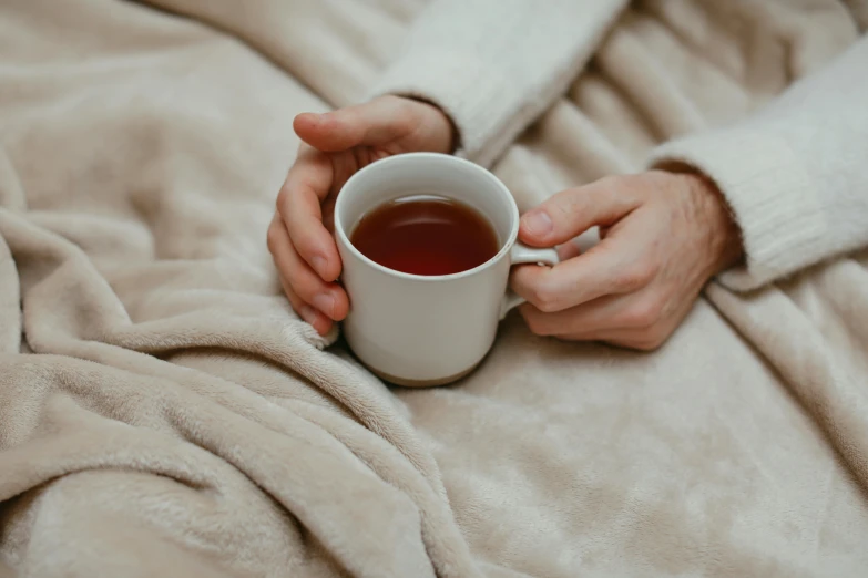 a person holding a cup of tea on a bed, trending on pexels, he is wearing a brown sweater, manuka, slightly red, curled up under the covers