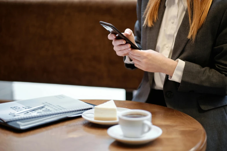 a woman sitting at a table looking at her cell phone, trending on pexels, girl in a suit, sitting on a mocha-colored table, thumbnail, corporate phone app icon