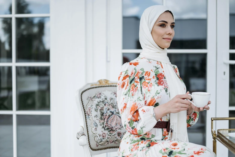 a woman sitting on a chair holding a cup of coffee, a portrait, inspired by Maryam Hashemi, trending on pexels, hurufiyya, wearing floral chiton, wearing ivory colour dress, muslim, standing outside a house