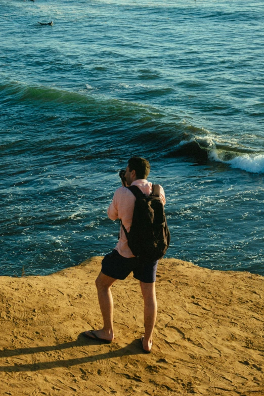 a man standing on top of a sandy beach next to the ocean, taking a picture, a man wearing a backpack, hugging, large waves hitting the cliff