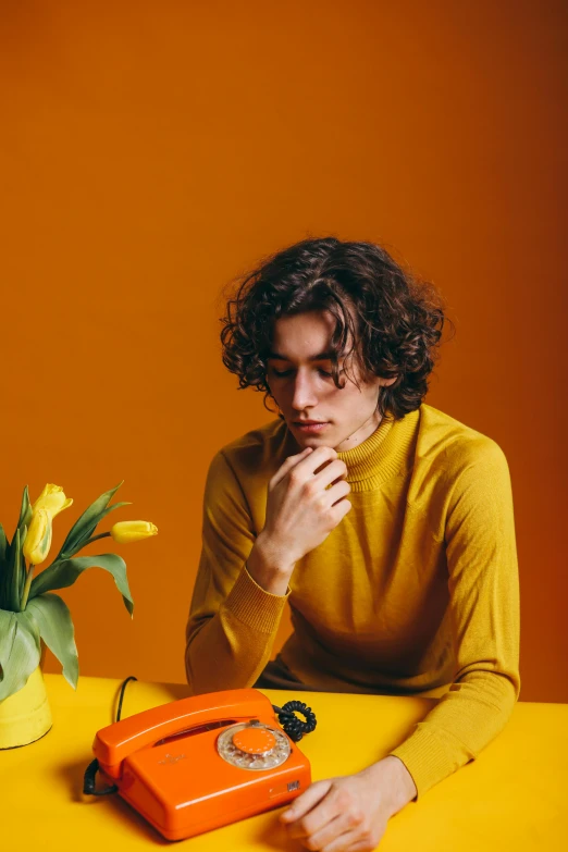 a man sitting at a table with an orange phone, an album cover, trending on pexels, wavy hair yellow theme, thinking pose, flowers, low quality photo