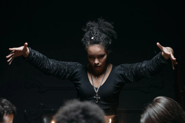 a woman standing in front of a group of people, an album cover, pexels contest winner, black arts movement, casting a protection spell, dark lighting, hands pressed together in bow, occult jewelry