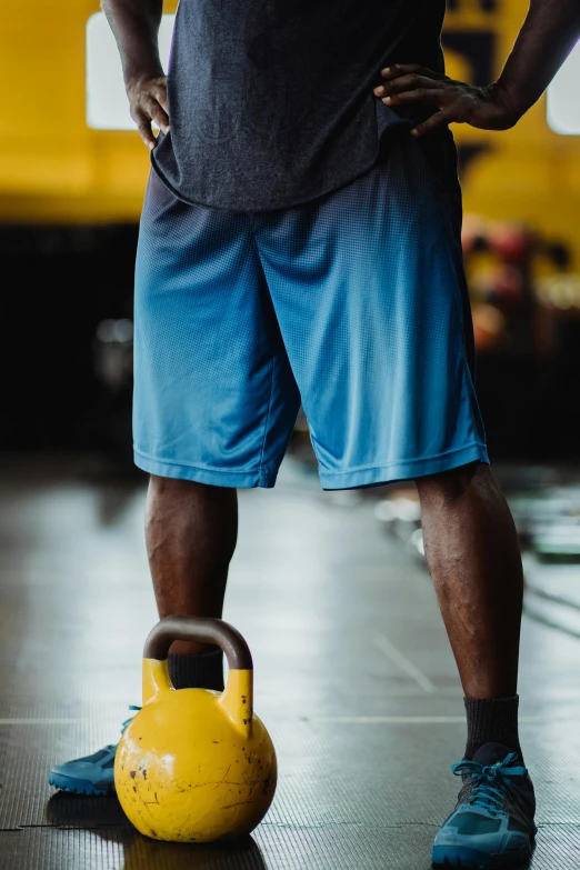 a man standing next to a kettlebell in a gym, by Samuel Washington Weis, pexels contest winner, happening, in blue and yellow clothes, human back legs and sneakers, athletic shorts, game ready