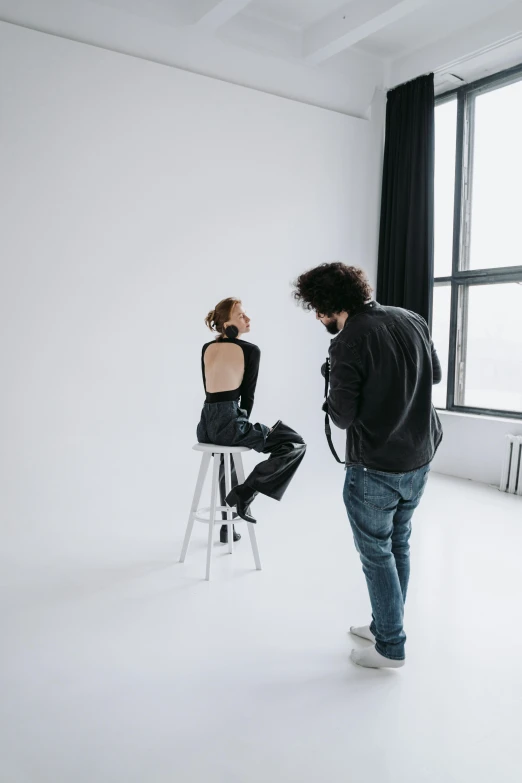 a man standing next to a woman in a white room, a picture, by Jonas De Ro, trending on unsplash, sitting on a stool, behind the scenes photo, model is wearing techtical vest, backshot