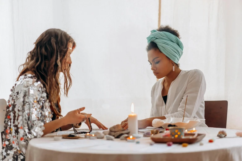 two women sitting at a table with candles, trending on pexels, process art, cloth head wrap, painted nails, in a white boho style studio, casting spells