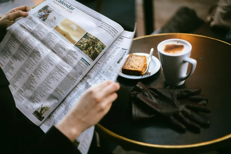 a person reading a newspaper next to a cup of coffee, by Daniel Gelon, trending on unsplash, sitting on a mocha-colored table, france, australian, daily specials