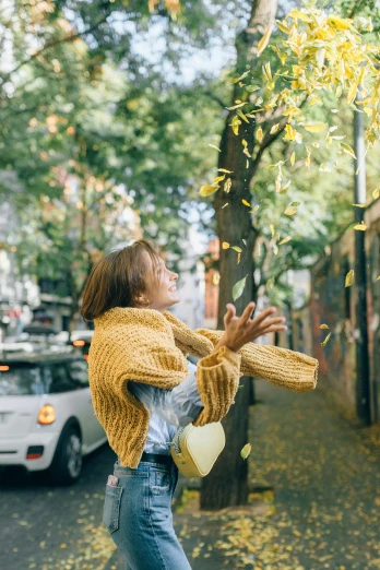 a woman throwing leaves in the air on a city street, pexels contest winner, wearing casual sweater, yellow theme, looking around a corner, loosely cropped