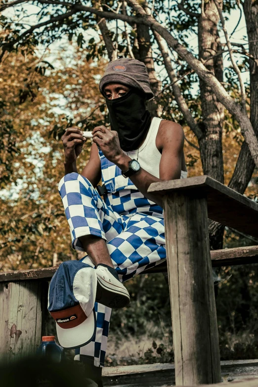 a man sitting on top of a wooden bench, an album cover, inspired by Ras Akyem, trending on pexels, visual art, blue checkerboard dress, balaclava, overalls, ( ( theatrical ) )