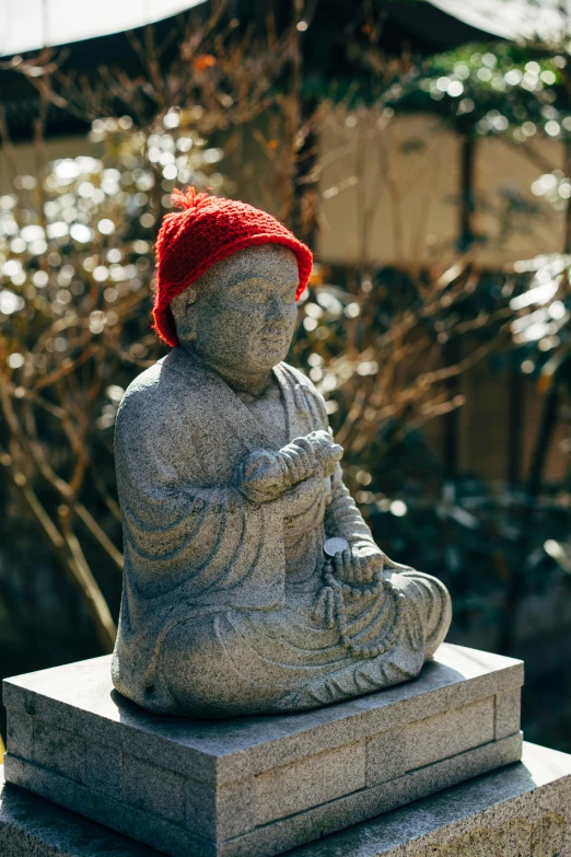 a statue of a person wearing a red hat, a statue, inspired by Tani Bunchō, unsplash, buddha, sitting in the garden, winter sun, grey