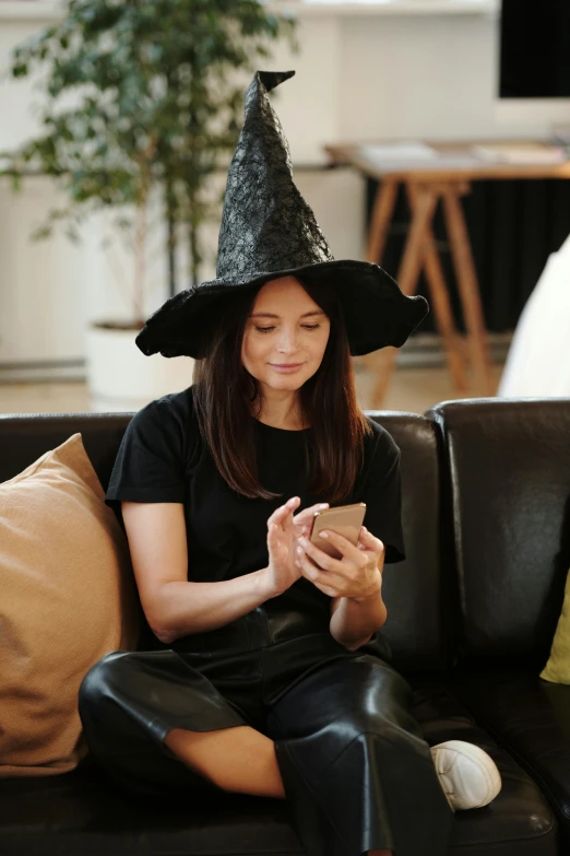 a woman in a witch hat sitting on a couch, trending on pexels, checking her phone, gemma chan, costume, ( 3 1