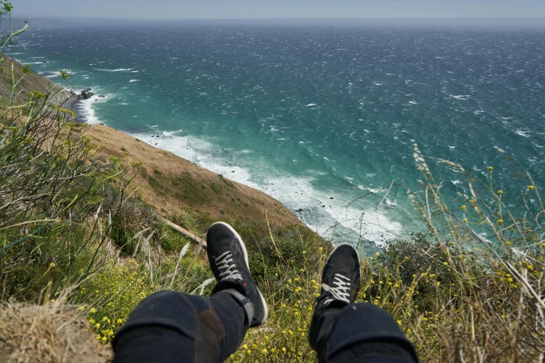 a person sitting on top of a hill next to the ocean, happening, sneaker photo, california coast, vertigo - inducing, profile image