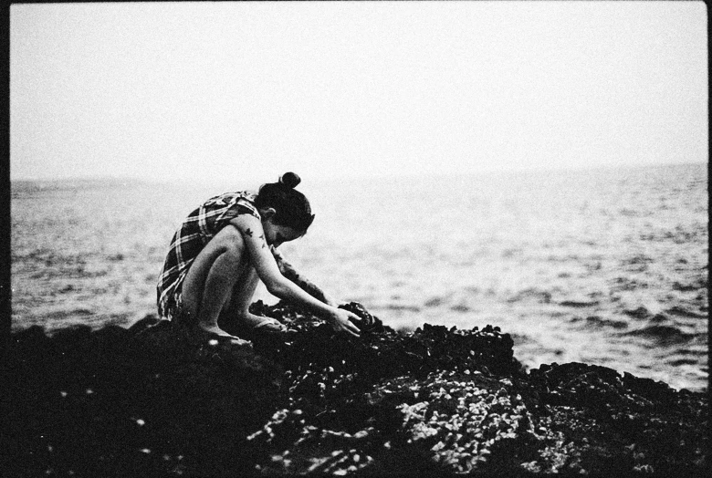 a woman sitting on top of a rock next to the ocean, a black and white photo, digging, jovana rikalo, grainy low quality, hands