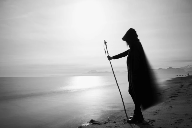 a black and white photo of a person on the beach, a black and white photo, unsplash, romanticism, wizard magic staff, holding a spear, wearing cloak and hood, siluette