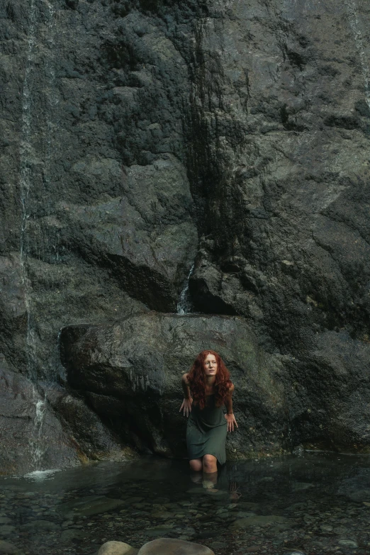 a woman standing in the water in front of a waterfall, by Elsa Bleda, renaissance, a redheaded young woman, with lots of dark grey rocks, ignant, low quality photo