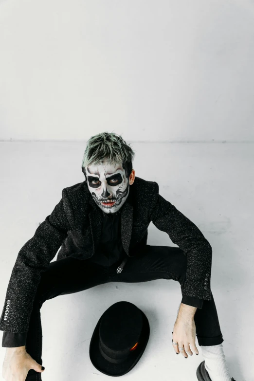 a man sitting on top of a white floor next to a hat, an album cover, inspired by Taro Yamamoto, trending on pexels, skull face paint, a silver haired mad, black mask, zombie face