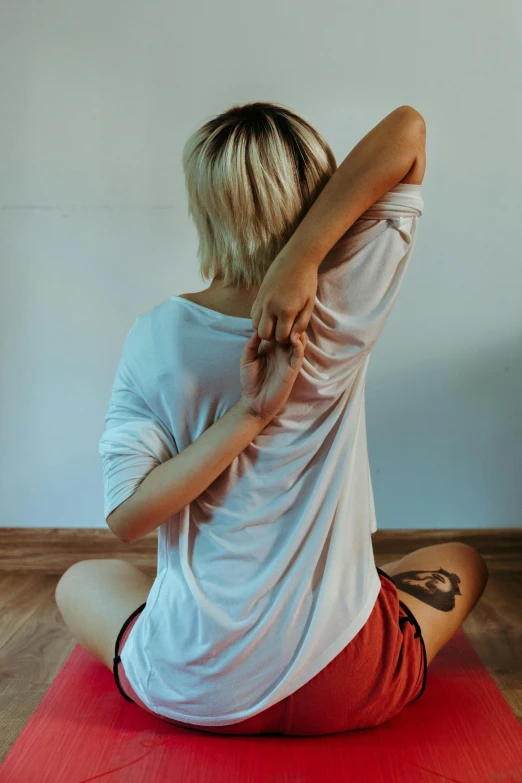 a woman sitting on top of a red yoga mat, a tattoo, unsplash, clothed in white shirt, showing her shoulder from back, blond, esthetic photo