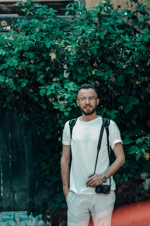 a man standing in front of a bush with a camera, by Adam Szentpétery, dressed in a white t-shirt, a man wearing a backpack, doug walker, ignant