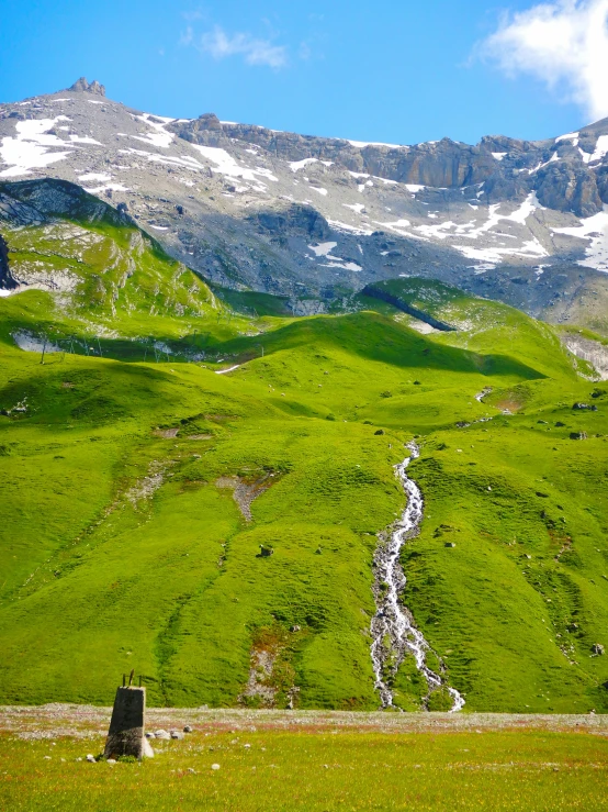 a grassy field with a mountain in the background, inspired by Werner Andermatt, pexels contest winner, les nabis, small flowing stream from wall, high view, high elevation, full of greenish liquid