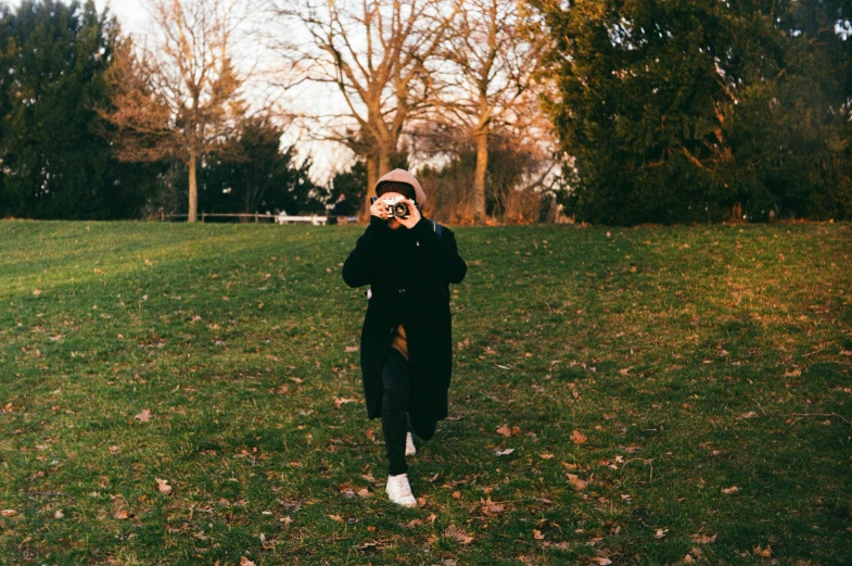 a woman standing on top of a lush green field, a picture, by Emma Andijewska, unsplash, visual art, taken on a 1960s kodak camera, walking at the park, during autumn, holding a dslr camera