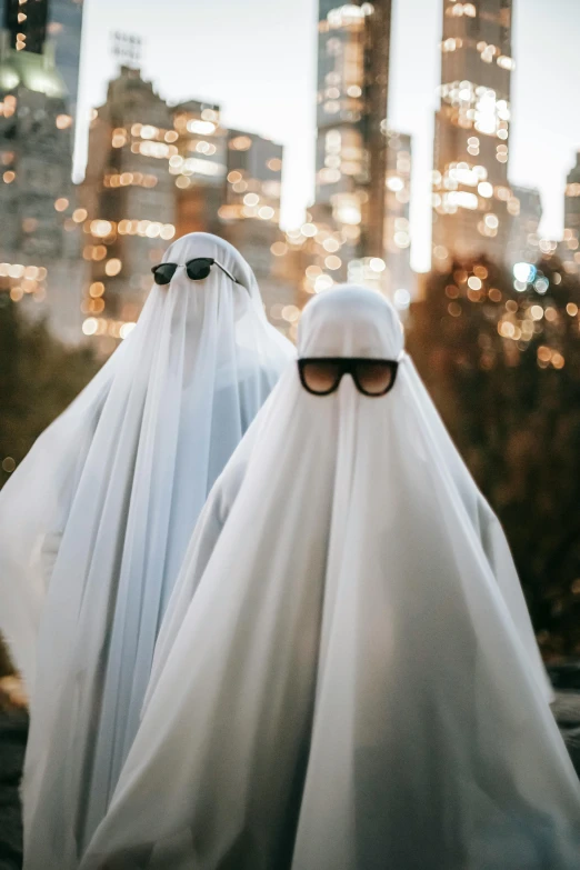 two people dressed in white standing in front of a cityscape, an album cover, trending on unsplash, happening, halloween ghost under a sheet, aviators, 2019 trending photo, veiled