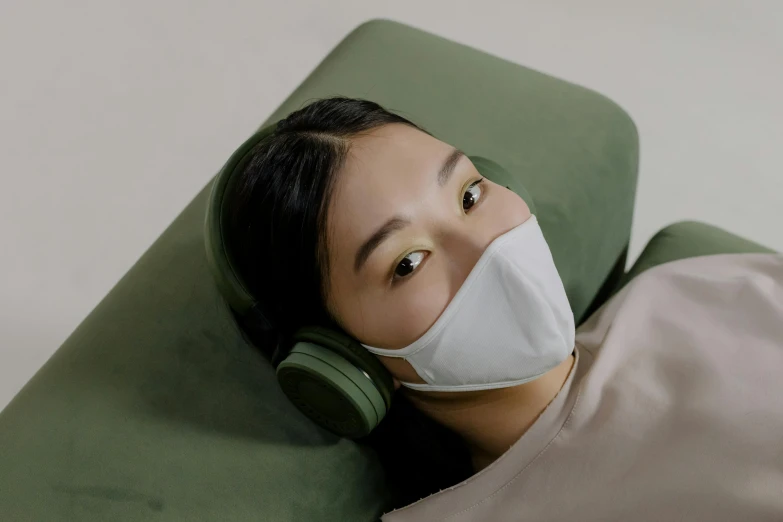 a woman laying on a green couch wearing a face mask, inspired by Fei Danxu, trending on pexels, sōsaku hanga, with head phones, haze over the shoulder shot, designer product, hong june hyung