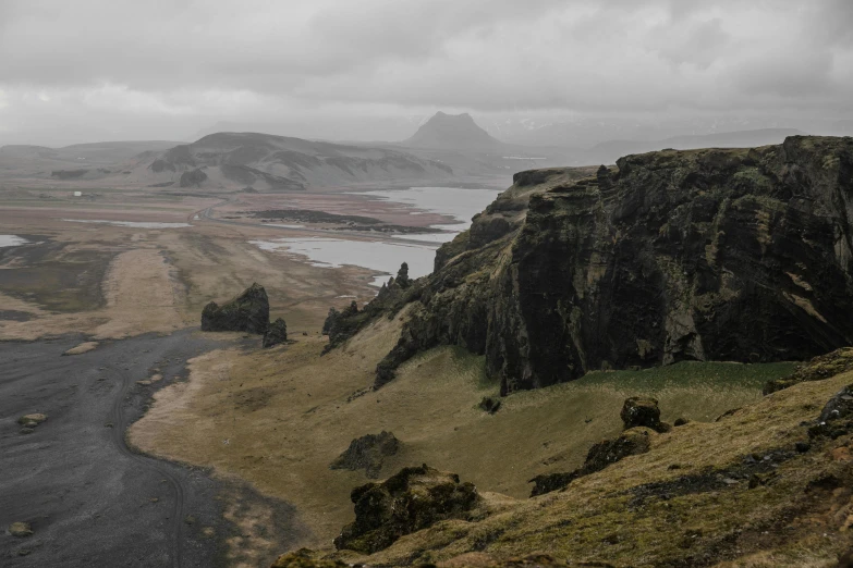 a man standing on top of a mountain next to a river, inspired by Þórarinn B. Þorláksson, pexels contest winner, renaissance, overcast weather, black volcano afar, trees and cliffs, brown