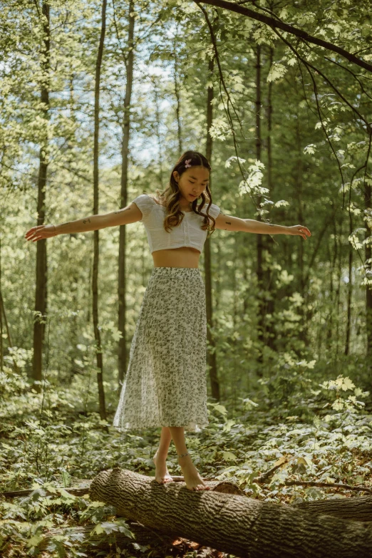 a woman standing on a log in the woods, by Dulah Marie Evans, pexels contest winner, renaissance, wearing crop top, waving, white shirt and green skirt, patterned clothing