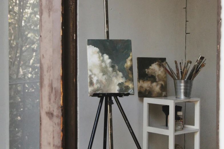 a painting that is on a tripod in a room, inspired by Vincenzo Irolli, unsplash, tonalism, clouds of smoke, easel, gray canvas, with clouds in the sky