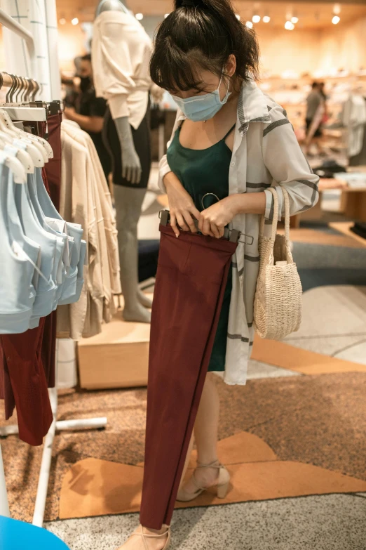 a woman standing in front of a rack of clothes, red shirt brown pants, wearing facemask, singapore, thigh focus