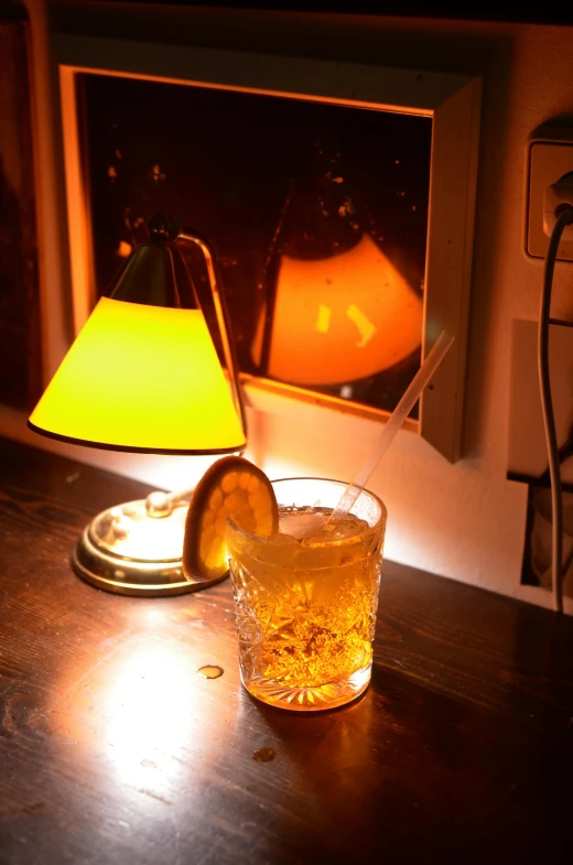 a drink sitting on top of a wooden table next to a lamp, renaissance, yellow light, light cone, old fashioned, ignant