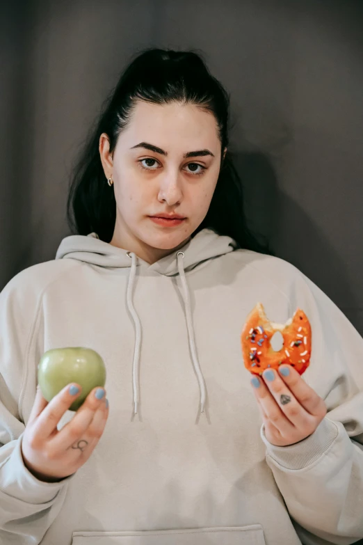 a woman holding an apple and a donut, a colorized photo, trending on pexels, wearing sweatshirt, intricate heterochromia sad, 👅 👅, charli xcx