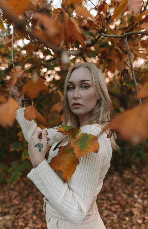 a woman standing under a tree with leaves, inspired by Elsa Bleda, trending on pexels, portrait of kim petras, white and orange, low quality photo, autum