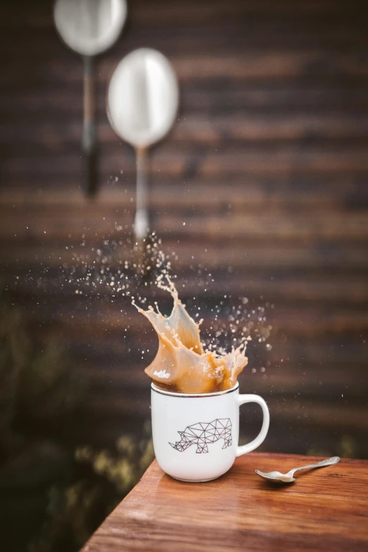 a spoon sitting on top of a wooden table next to a cup of coffee, splashing, flying pig, caramel. rugged, full product shot