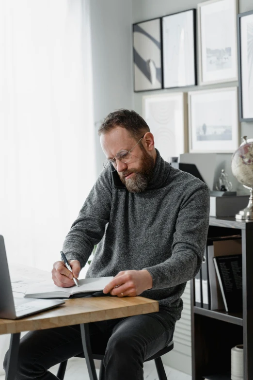 a man sitting at a desk writing on a piece of paper, online, grey, man with beard, home office