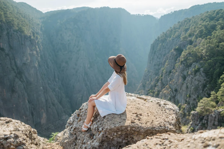 a woman sitting on a rock overlooking a valley, pexels contest winner, les nabis, wearing a white sundress, avatar image, sharp cliffs, wearing a straw hat and overalls