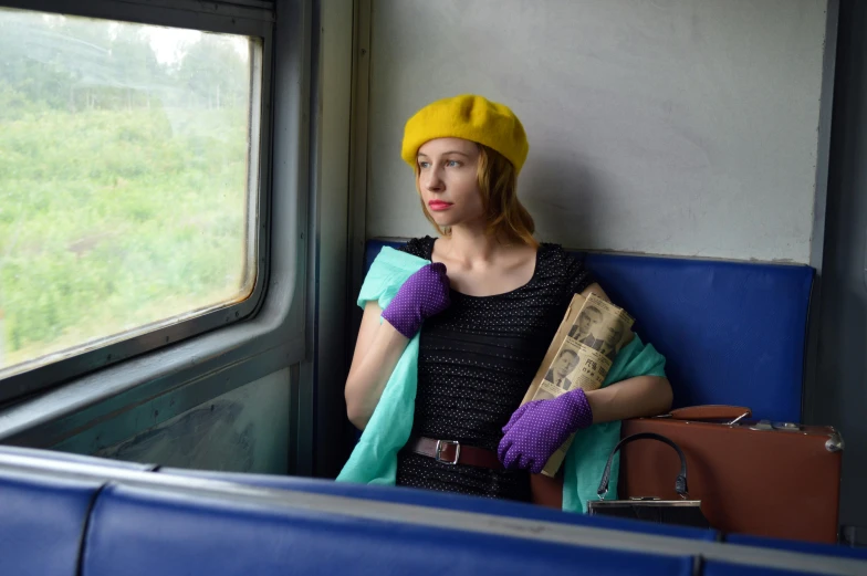 a woman sitting on a train looking out the window, inspired by Wes Anderson, regionalism, russian clothes, wearing a purple cap, wearing a yellow dress, wearing gloves