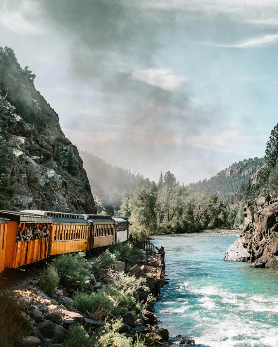 a train traveling down train tracks next to a river, a colorized photo, by Morgan Russell, unsplash contest winner, colorado mountains, photo for vogue, reddit post, smokey