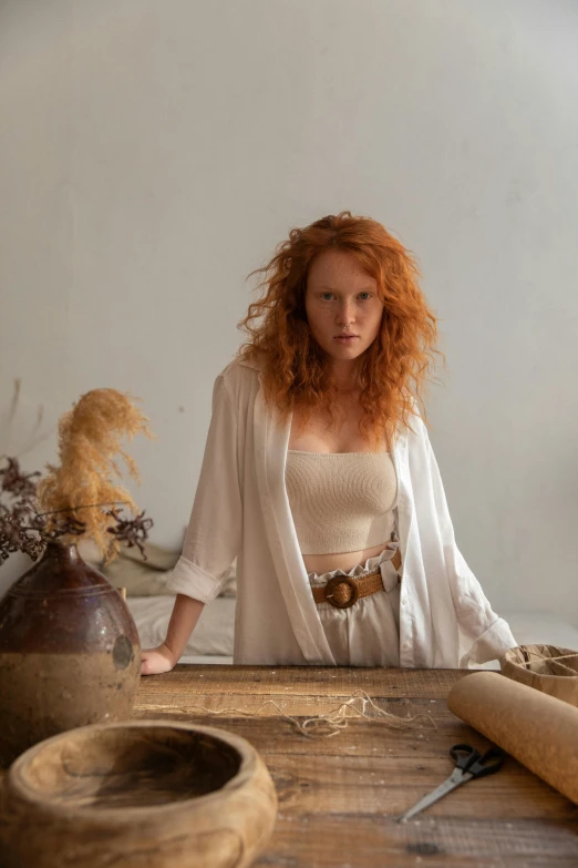 a woman that is standing in front of a table, inspired by Filippino Lippi, renaissance, wild ginger hair, in a white boho style studio, red dust, on location