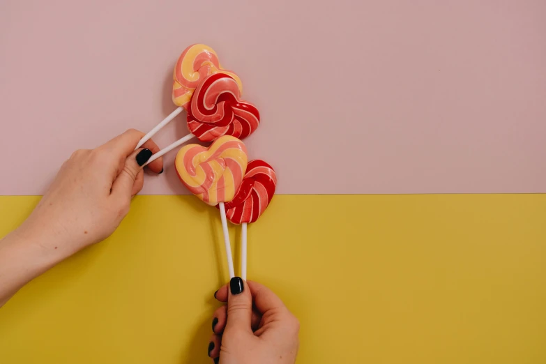 a close up of a person holding two lollipops, inspired by Ödön Márffy, trending on pexels, pink and yellow, red swirls, food styling, background image