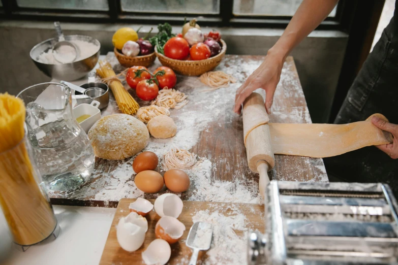 a person rolling out dough on a table, pexels contest winner, 💋 💄 👠 👗, the table is full of food, botanicals, pasta