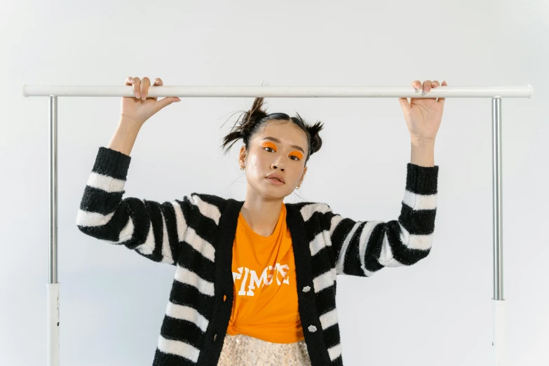 a woman in an orange shirt and a black and white striped cardigan, inspired by Kim Tschang Yeul, pexels contest winner, hanging from the ceiling, resembling a mix of grimes, ayami koj ima, pose 4 of 1 6