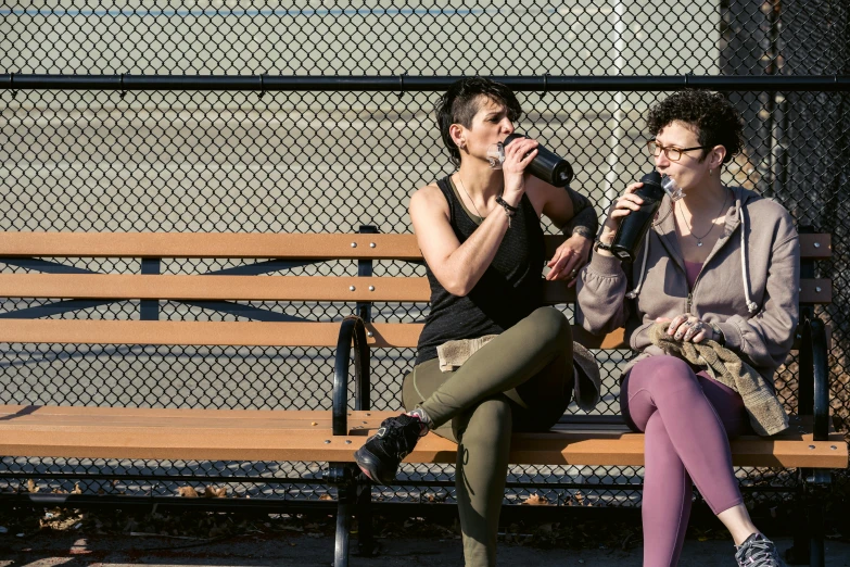 a couple of people that are sitting on a bench, by Nina Hamnett, pexels, happening, water bottles, lesbian, holding a drink, muscular and exhausted woman