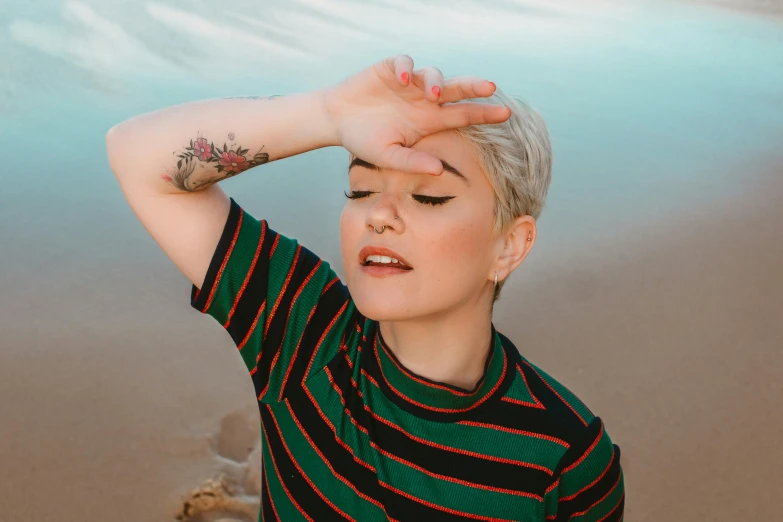 a woman standing on top of a beach next to a body of water, a tattoo, inspired by Elsa Bleda, trending on pexels, short platinum hair tomboy, half - closed eyes, wearing stripe shirt, photograph of a sleeve tattoo