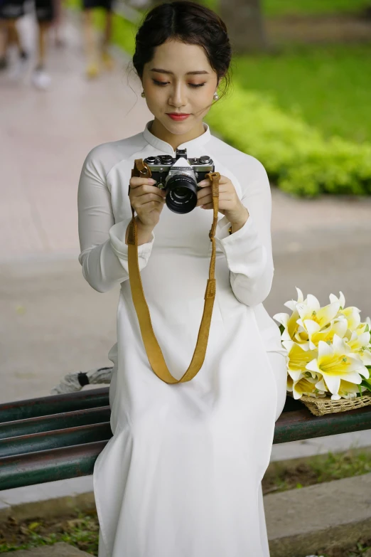 a woman sitting on a bench holding a camera, a picture, inspired by Ruth Jên, ao dai, white soft leather model, basic, with yellow flowers around it