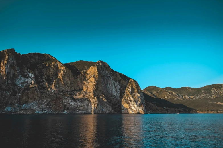 a body of water with a mountain in the background, by Carlo Martini, pexels contest winner, romanticism, coastal cliffs, late afternoon light, clear blue skies, thumbnail